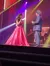 Two performers are engaging with each other on a brightly lit stage, with one wearing a striking red gown and the other in a suit.