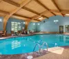 An indoor swimming pool with clear blue water is surrounded by multiple chairs and large windows allowing ample natural light all set under a vaulted wooden ceiling