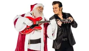 A person dressed as Santa Claus plays a red guitar while smiling at a performer styled like Elvis Presley who is also holding a guitar, against a white background.