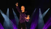 A person wearing a purple velvet shirt is posing dramatically with raised arms against a dark background with beams of blue and purple light.