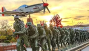 The image depicts a powerful sculpture installation featuring a group of soldier statues in historical military gear, advancing beneath a vintage fighter aircraft, against a vividly colored sunset sky.