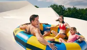 Three people are enjoying a sunny day at a water park, sliding down in a large inflatable ring with big smiles on their faces.