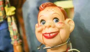 The image shows a close-up of a vintage-looking toy with a smiling, freckled face and blue eyes, accompanied by blurred background figures.