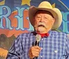 A person in a cowboy hat and bandana is speaking into a microphone against a colorful backdrop with the word Fiesta