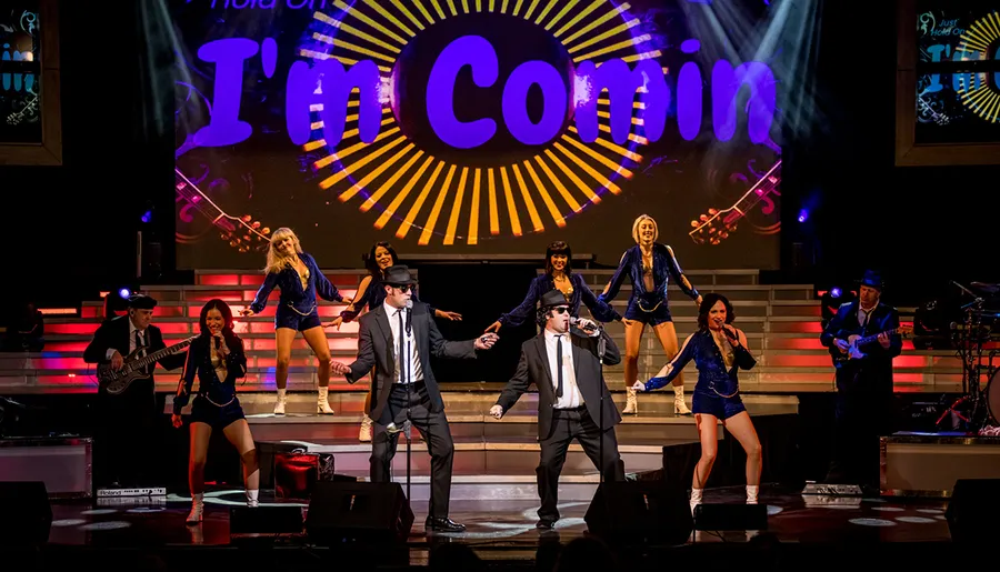 A vibrant live music performance featuring two central singers in suits and sunglasses, flanked by a band and dancers on a stage with colorful lighting and the words I'm Comin in the background.