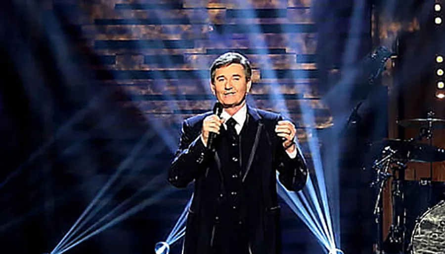 A person in a suit is performing on stage with a microphone, highlighted by dramatic blue stage lighting and a backdrop that gives the appearance of a starry night.