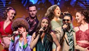 A group of performers in vibrant, retro-themed costumes are singing and dancing on stage with a dynamic backdrop that suggests a lively musical atmosphere.