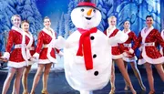 A person in a snowman costume is surrounded by smiling dancers in festive outfits on a winter-themed stage.