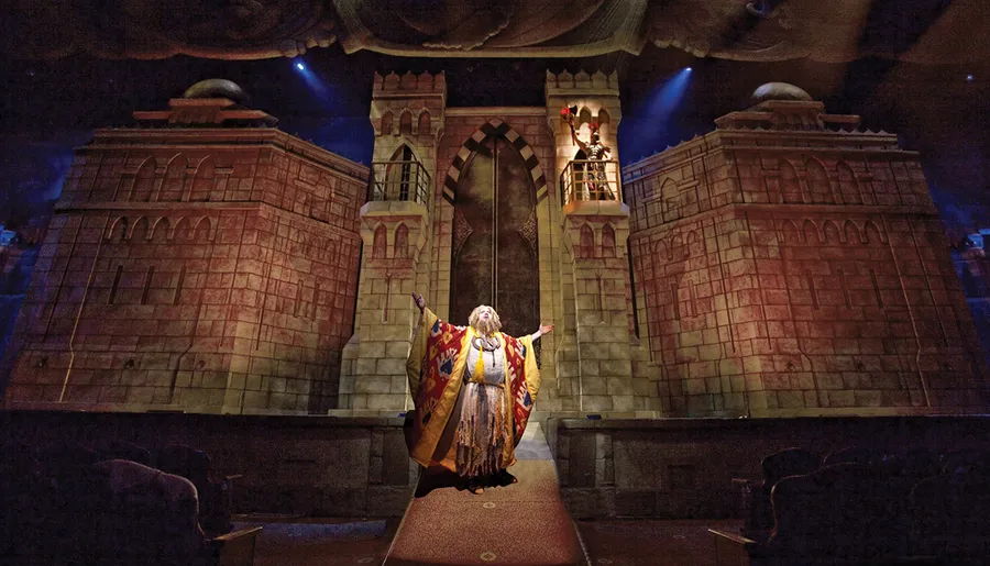 A performer dressed in lavish, medieval-style robes is standing with outstretched arms on a theater stage set to resemble a grand castle gate.