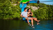 A man and a woman are smiling and enjoying a chairlift ride over a body of water, surrounded by lush greenery.