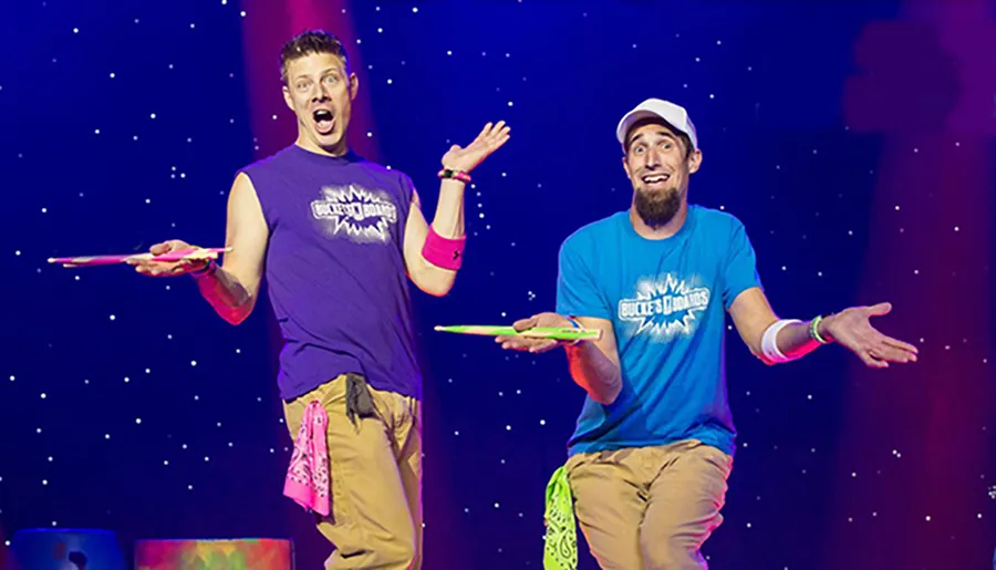 Two performers are on stage with surprised expressions, holding spinning plates on sticks against a starry background.