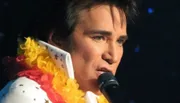 A person is performing onstage with a microphone, dressed in an Elvis Presley-inspired outfit adorned with a colorful lei.