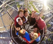 A group of cheerful young people is posing for a fisheye lens photo from an elevated viewpoint on a sunny day.