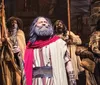 The image depicts actors in historical or biblical costumes performing in a theatrical production with one prominently featured man in a red shawl and beard looking upwards