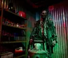 The image shows a horror-themed figure possibly a zombie standing next to a shelf filled with jars and a gas can in a dimly lit room with corrugated metal walls