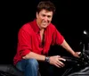 A man in a red shirt smiles while sitting on a motorcycle against a black background