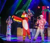 A person in a colorful parrot costume with a pirate hat dances on a stage next to a man singing into a microphone with other performers in tropical attire and illuminated palm trees in the background