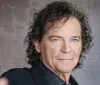The image shows a confident middle-aged man with curly hair wearing a black blazer posing for the camera against a textured grey background