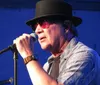 A musician wearing sunglasses and a hat performs into a microphone with musical equipment in the background