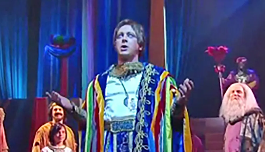 A performer in a colorful costume appears to be singing or acting passionately on stage, surrounded by other characters in elaborate attire, suggesting a theatrical production.