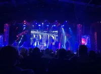 The image shows a theatrical performance underway, with actors on stage under colorful stage lighting, in front of an audience.