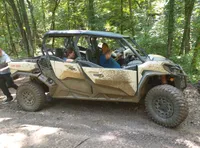 A mud-spattered off-road vehicle is surrounded by greenery, with two people inside and another person standing by it, indicating a break during an off-roading adventure.