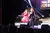 Two performers in vibrant costumes are singing onstage while a band plays musical instruments in the background during a live concert.