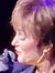 A performer is singing on stage with colorful lighting and band members in the background.