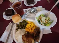 This image shows a hearty meal consisting of a dinner roll, green beans, macaroni and cheese, gravy-covered meat with mashed potatoes, a piece of fried chicken, salad on a small plate, a slice of chocolate cake, and a glass of iced tea, all arranged on a maroon-colored table.