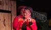 A person in a red jacket with gold trimmings and a black hat is performing on stage with a microphone, making a gesture, while another individual stands behind on a set resembling a wooden structure, possibly during a theatrical or entertainment show.
