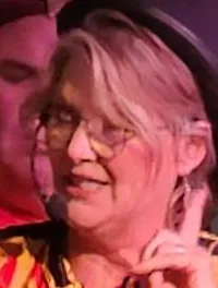 A person in a red jacket with gold trimmings and a black hat is performing on stage with a microphone, making a gesture, while another individual stands behind on a set resembling a wooden structure, possibly during a theatrical or entertainment show.