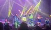 A group of performers in brightly colored costumes is singing and dancing on a lit stage in front of an audience.