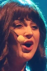 A person in a red top and beige fringed skirt is singing on stage with a microphone headset, illuminated by stage lighting.