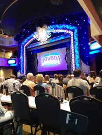 An audience is seated at tables in a theater, waiting for the performance of a show titled