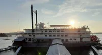 The image shows a moored paddle-wheel steamboat at sunset with the sun low in the sky behind it, casting a warm glow over the scene.
