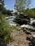 The image depicts a serene artificial river with cascading waters flanked by rocky banks, lush greenery, and rustic structures, possibly a part of an amusement park or themed attraction.