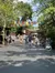The image displays a bustling theme park pathway lined with trees and festooned with colorful banners, with visitors strolling, some pushing strollers, under the bright and sunny sky.