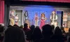 An audience watches performers on stage engaged in a scene from a play, which appears to have a rustic or western theme.