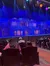The image depicts a theater auditorium with an ornately decorated stage set for a production, with audience members seated and waiting for the performance to begin under a lighting setup that casts a blue hue over the scene.