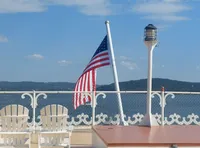 An American flag flutters in the breeze on a ferry deck with white chairs and a lamp post, against a backdrop of a calm lake and hills under a blue sky.