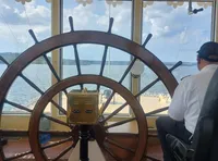 A person is at the helm of a boat, steering with a large wooden ship's wheel with a scenic view of the water and landscape ahead.