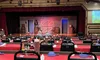 The image shows a dinner theater setup with numbered tables, black chairs, red tablecloths, and a stage with a rustic set design and lighting equipment overhead.