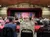 An audience sits at tables in a theater watching a stage with a closed red curtain and a set design visible on the side.