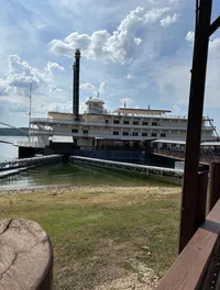 The image presents a large, multi-deck riverboat with iconic paddlewheel elements, docked beside a grassy waterfront under a partly cloudy sky.