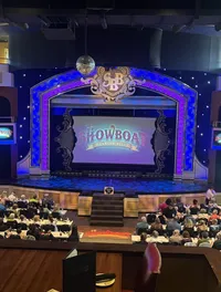 The image shows an ornate theater with a crowd of people seated at tables, facing a stage with a