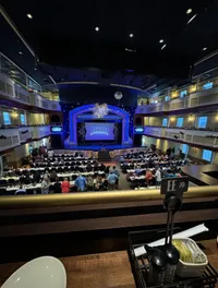 The image showcases an audience seated at tables in a multi-tiered theater awaiting the start of a performance, visible from a balcony perspective with a condiment caddy in the foreground.