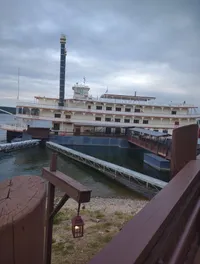 A riverboat with multiple decks is docked beside a wooden walkway with a quaint lantern in the foreground, against a backdrop of overcast skies.