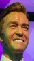 A wax figure of a smiling man dressed in a stylish suit stands against a purple geometric background.