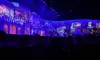 The image shows an audience watching a projection-mapped show or performance, featuring vibrant blue lighting and intricate architectural imagery.