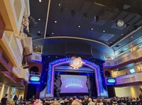 The image depicts the interior of a lavish theater with an audience waiting for a performance beneath a sign reading